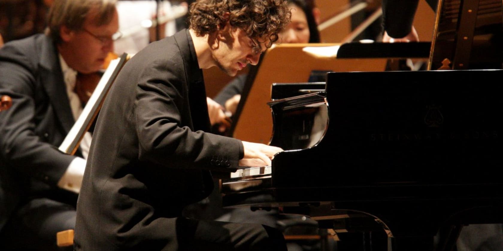 A man playing the piano during a concert, with other musicians in the background.