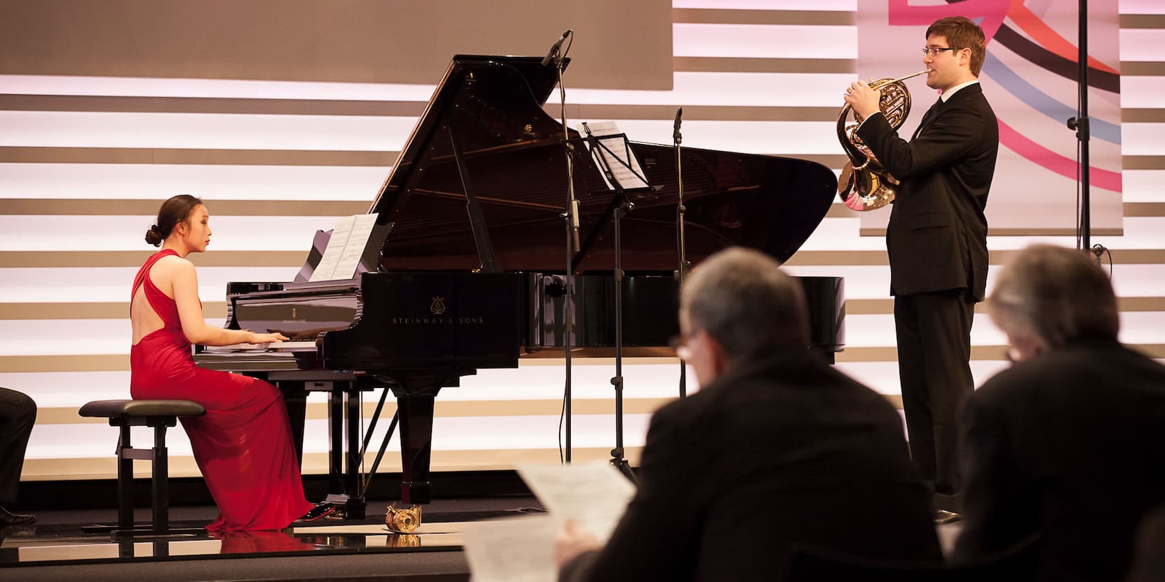 A woman in a red dress plays the piano while a man in a suit plays a horn. People are seated in the foreground, listening.