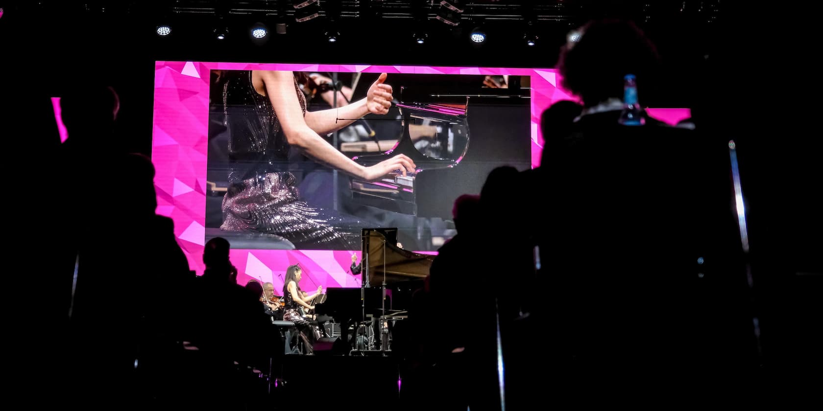 A concert with a pianist on stage, projected onto a large screen in the background. The audience sits in the dark.