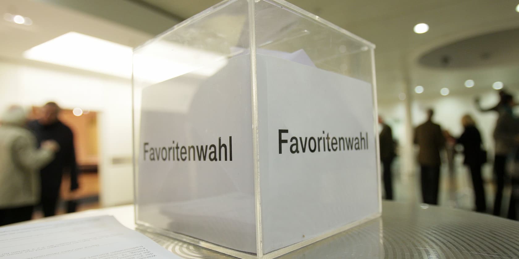 Ballot box labeled 'Favoritenwahl' with people in a room in the background.