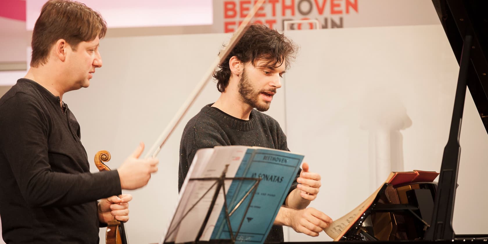 Two men practice music together; one is playing the violin and the other is at the piano. The Beethoven Festival logo is visible in the background.