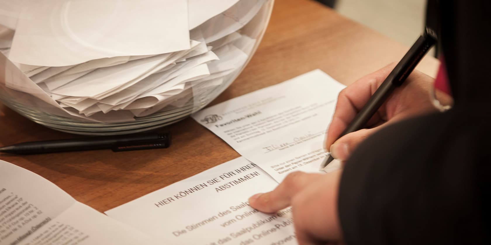 Person filling out a form next to a glass bowl filled with paper sheets.