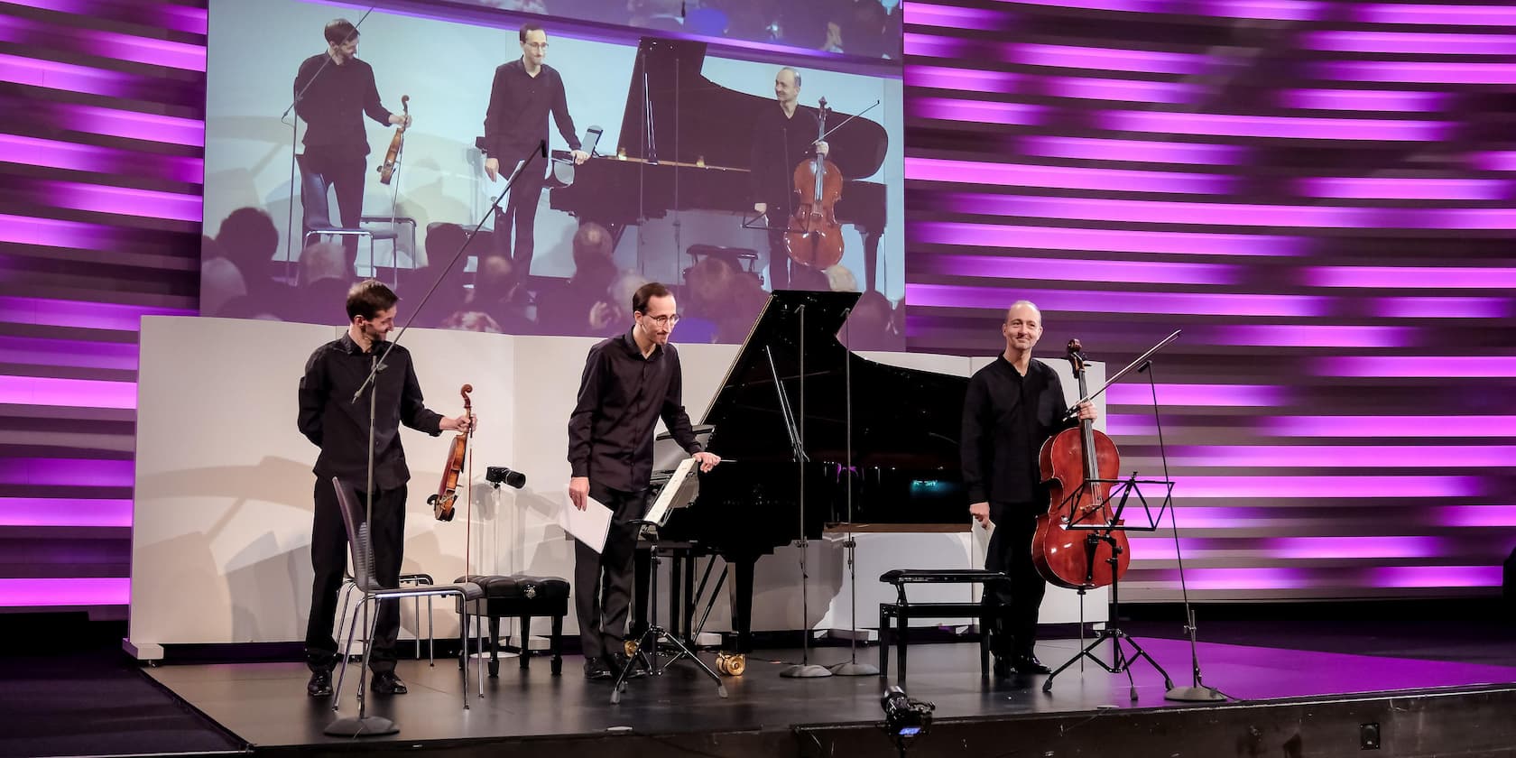 Three musicians on stage with a piano and a purple-lit background, during a performance.