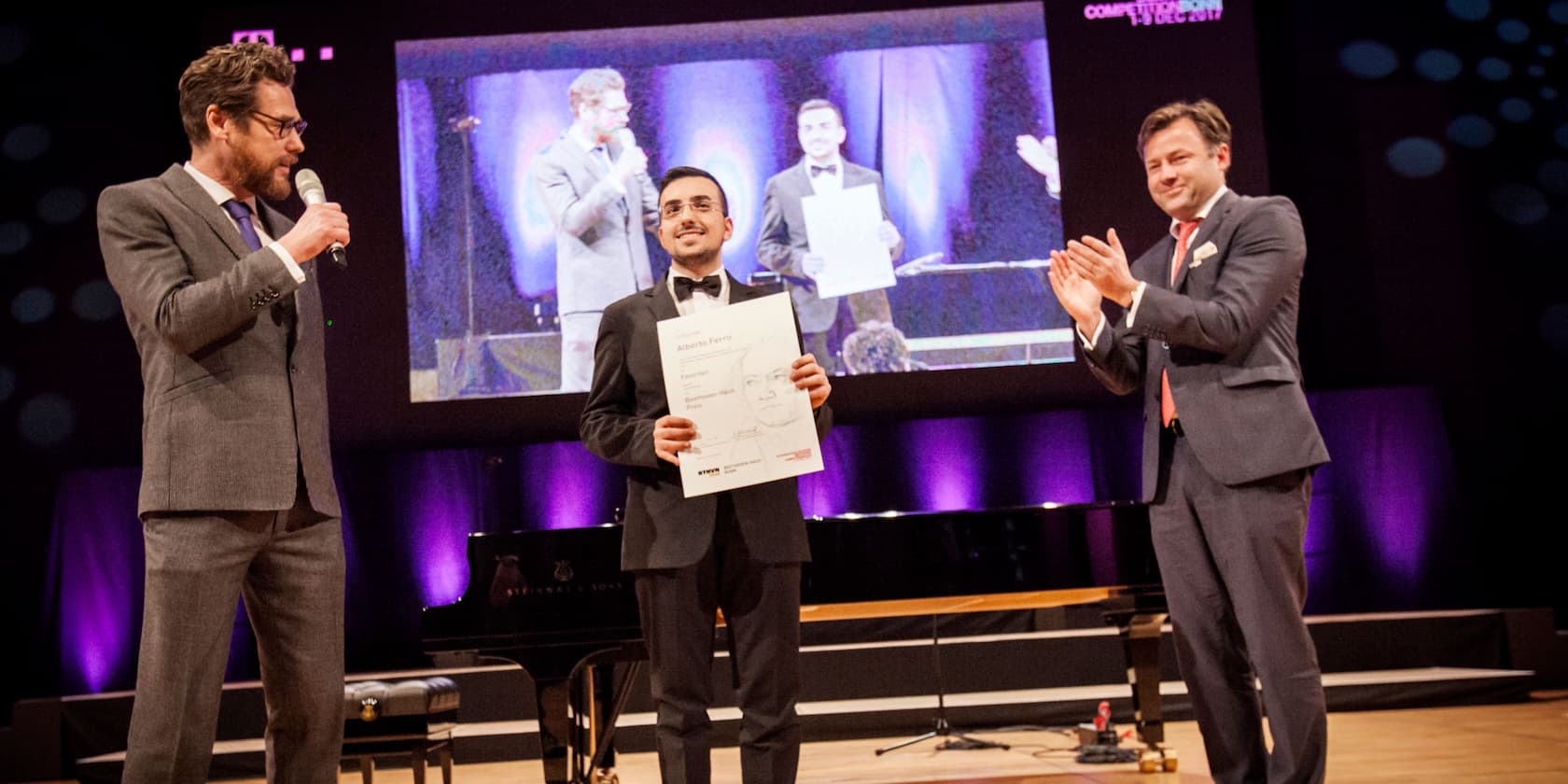 Three men in suits on a stage at an awards ceremony, one holding a certificate, another clapping.