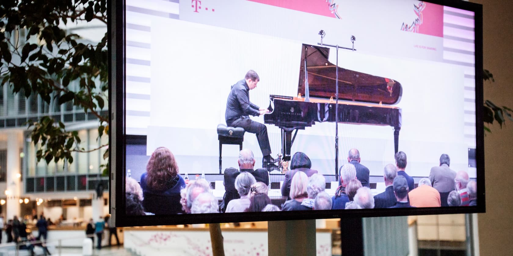 Ein Pianist spielt während eines Auftritts Klavier. Das Bild wird auf einem großen Bildschirm in einer Halle gezeigt. Ein Publikum schaut zu.