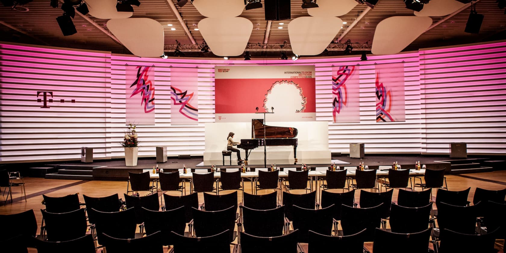 A woman plays the piano on a stage in a conference hall with empty rows of chairs in front of the stage. The background features the Telekom logo and colorful artworks.