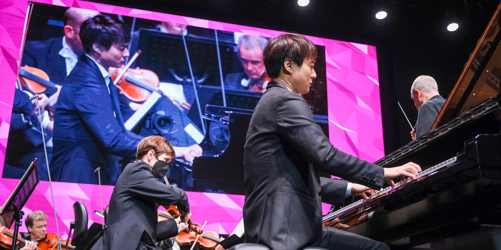 A pianist plays the piano on stage with an orchestra, while a projection of the performance is seen in the background.