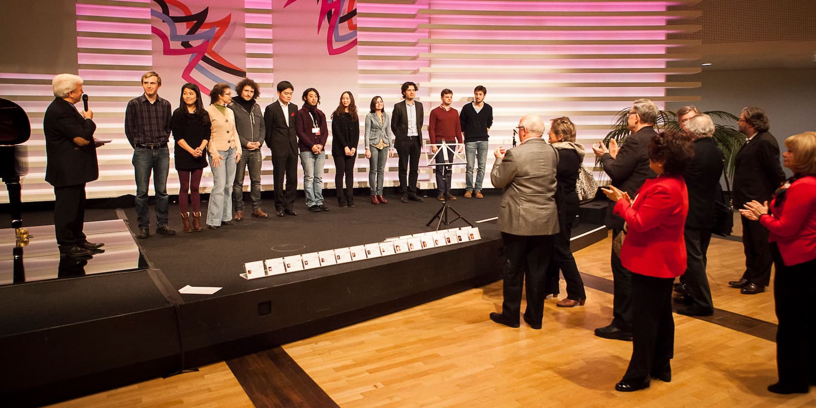 A group of ten people stands on a stage being introduced by a man with white hair. An audience applauds.