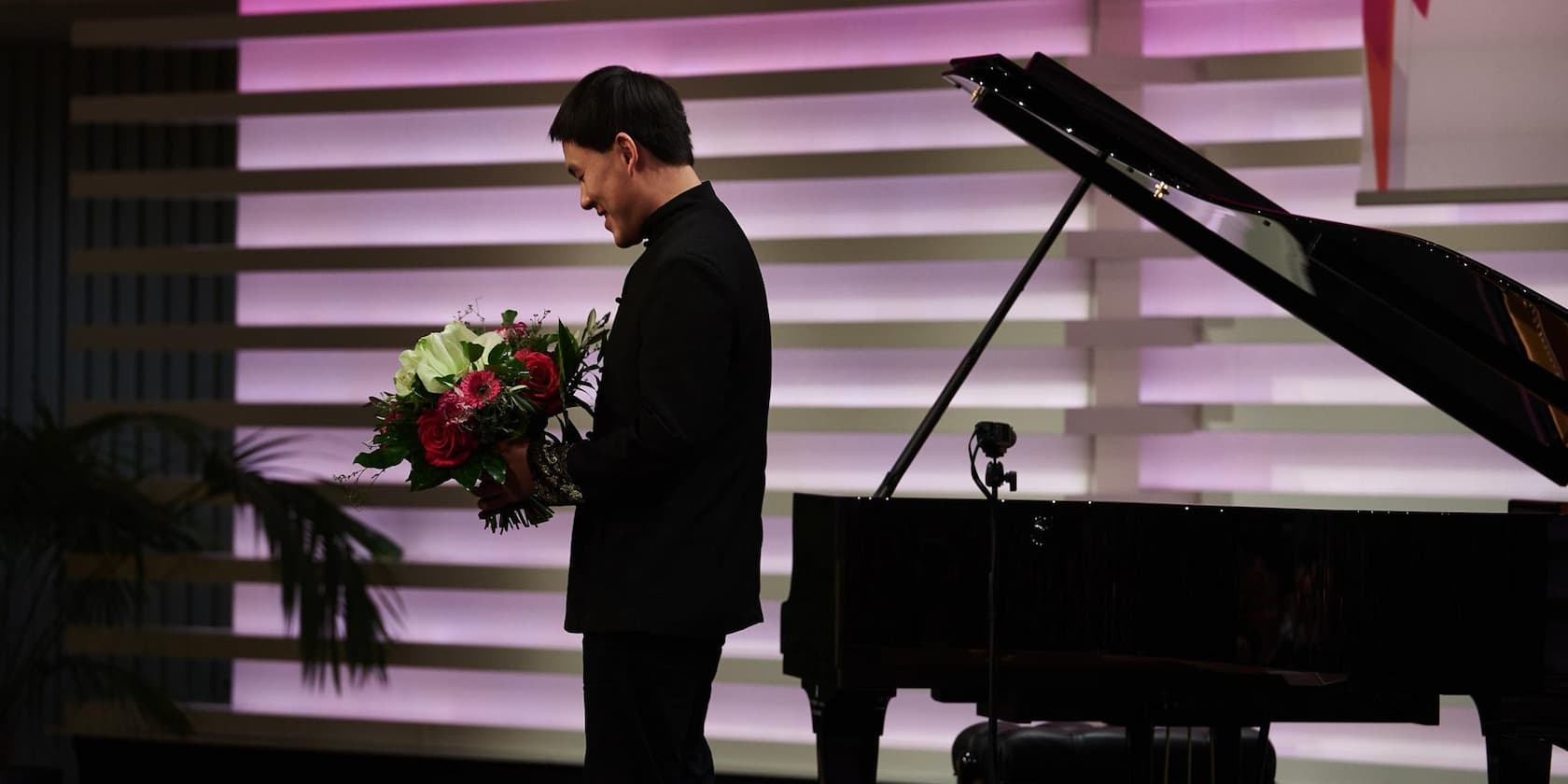 A man in a black suit jacket is holding a bouquet of flowers in front of a grand piano, with a background of horizontal lines lit in purple.