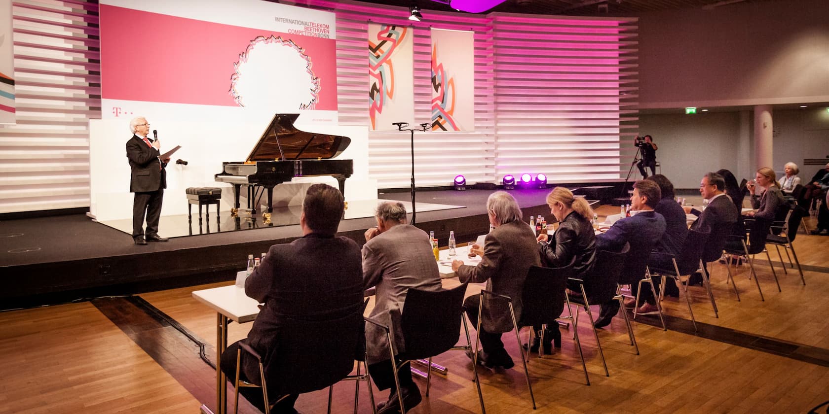 Man giving a speech at the International Telekom Beethoven Competition in Bonn, standing on a stage with a grand piano, while several audience members listen.