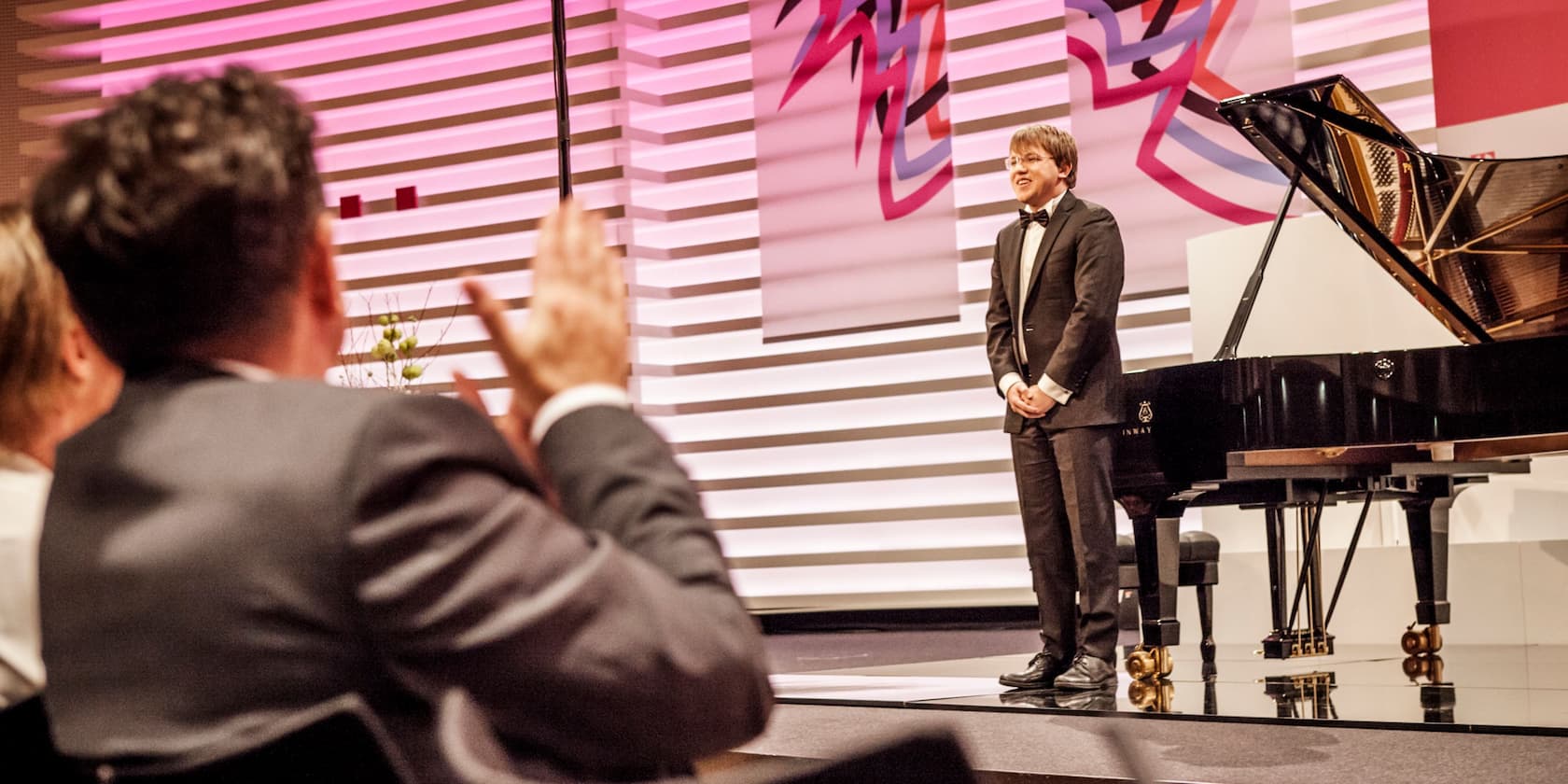 A man in a suit stands on a stage next to a grand piano, receiving applause from the audience.