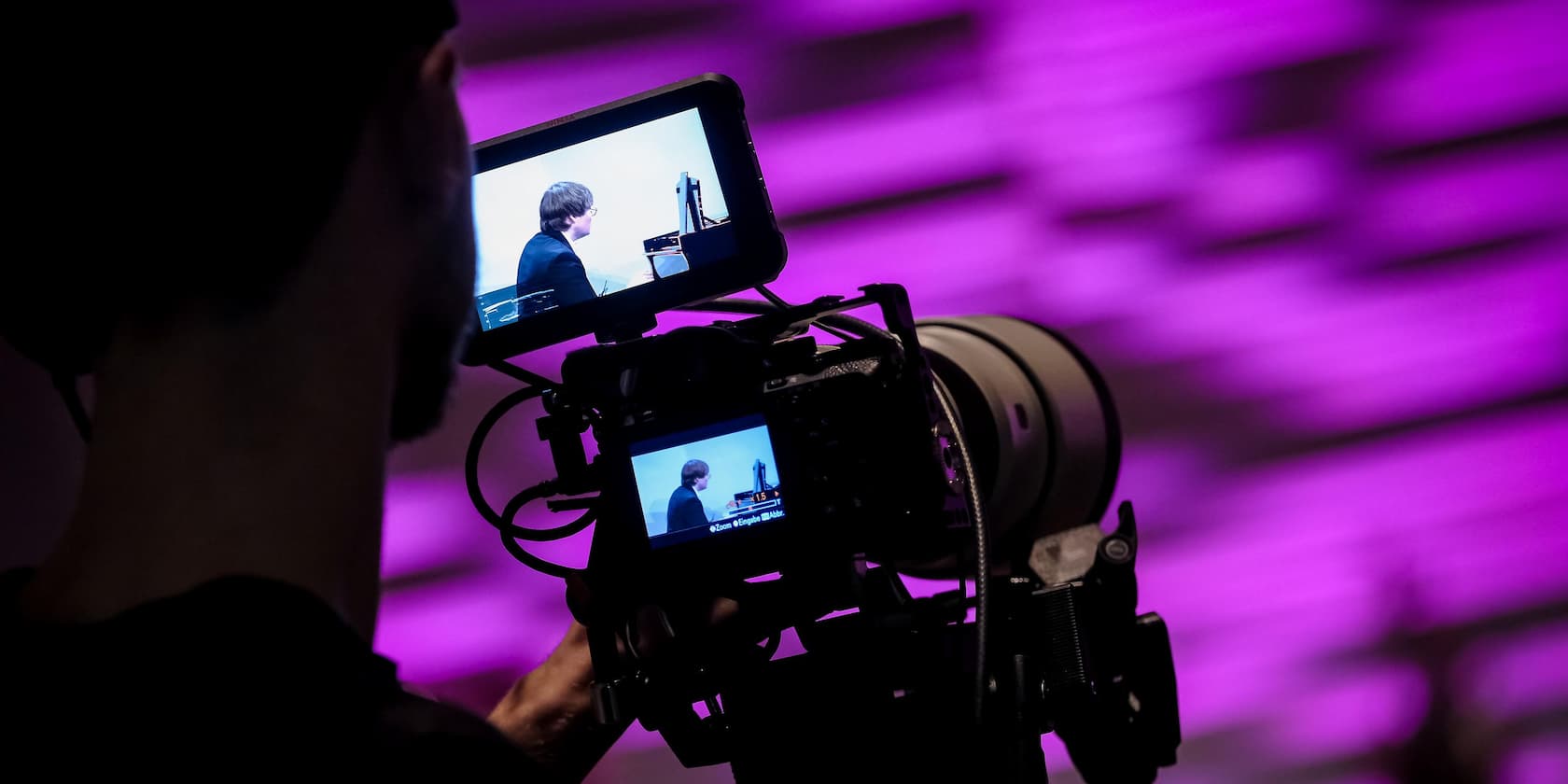 Cameraman filming a person playing the piano displayed on a screen, in front of a purple background.