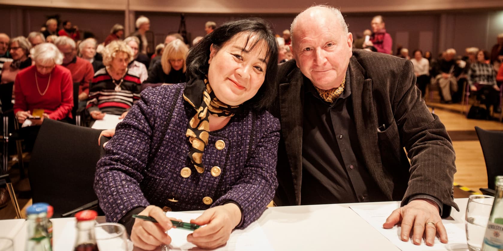 Two people sit at a table and smile, in a crowded hall.