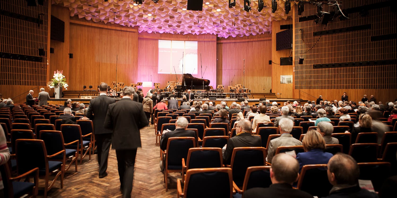 A concert hall with a view of the stage where a grand piano is placed. The audience is seated or moving around.