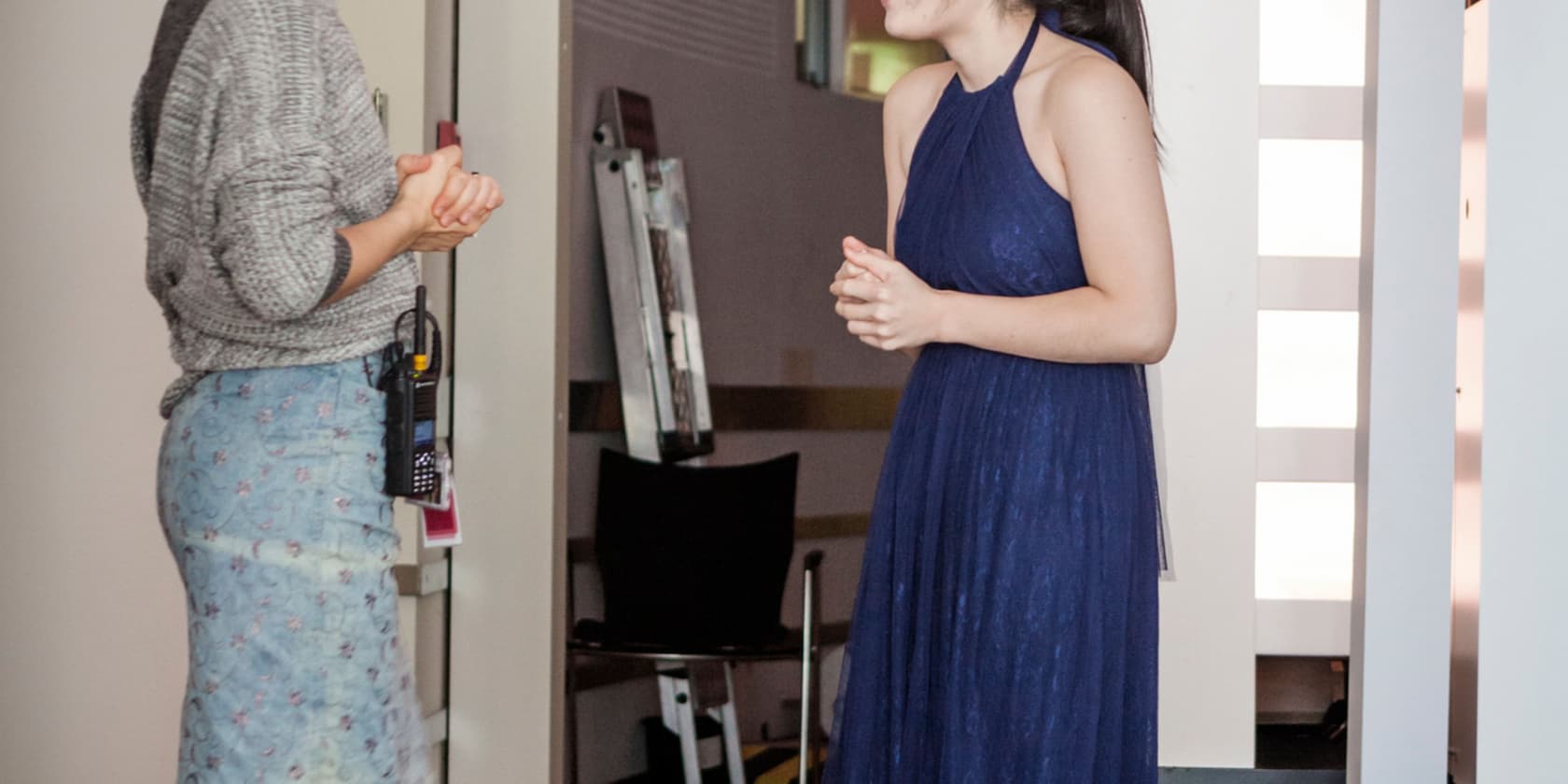 Two women are talking; one is wearing a blue dress, the other is casually dressed and has a walkie-talkie on her hip.