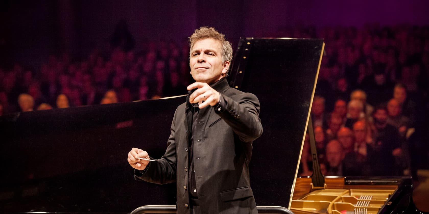 Conductor leading an orchestra in a concert hall, with an audience in the background.