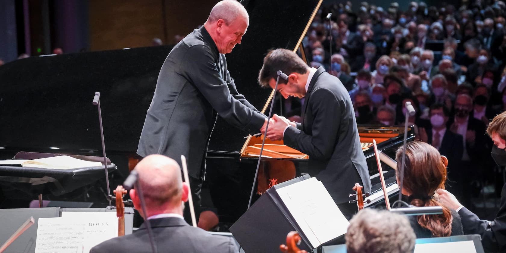 A conductor and a pianist shake hands in front of a grand piano, with an orchestra and audience in the background.