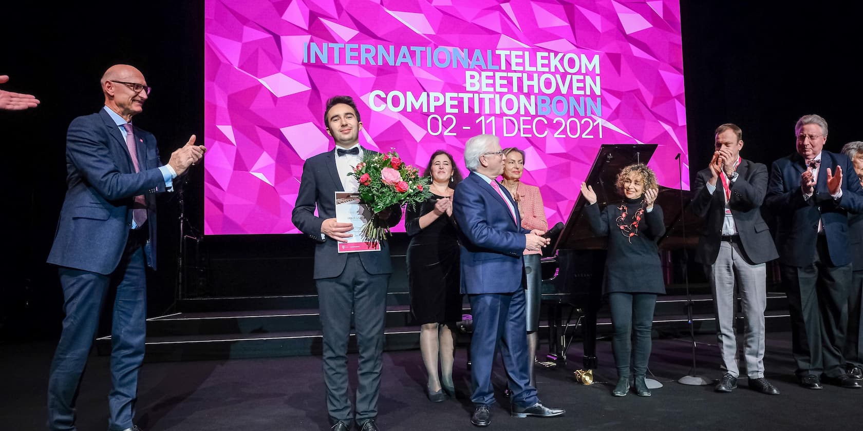 Gewinner der International Telekom Beethoven Competition Bonn 2021 mit Blumenstrauß und Urkunde vor einer Gruppe applaudierender Menschen.