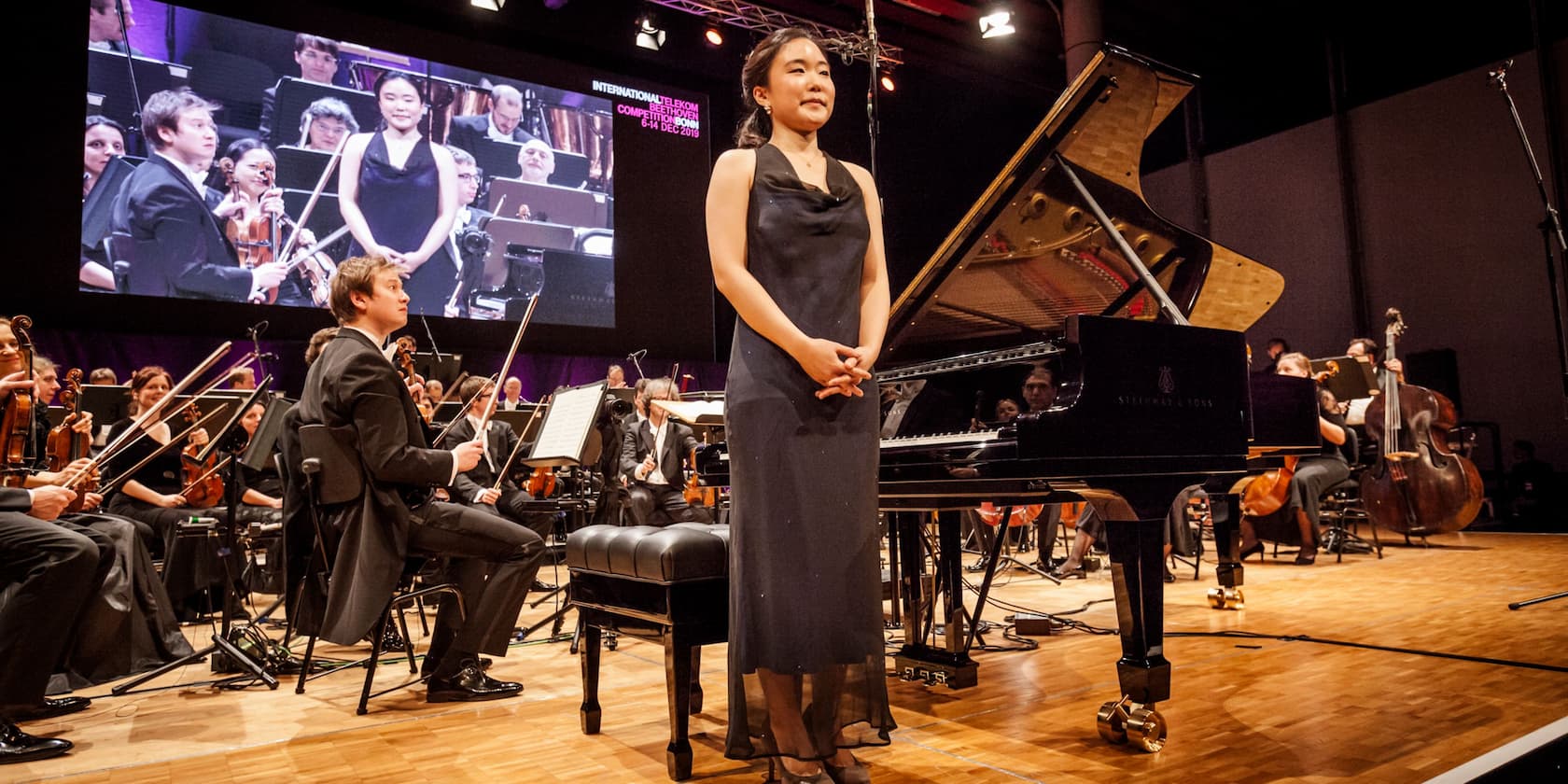 Pianist steht vor einem Flügel auf einer Konzertbühne, umgeben von einem Orchester, mit einem Projiziertem Bild im Hintergrund, das „International Telekom Beethoven Competition 6-14 Dec 2019“ anzeigt.