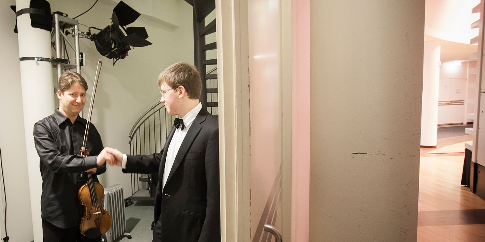 Two men, one holding a violin, shaking hands backstage.