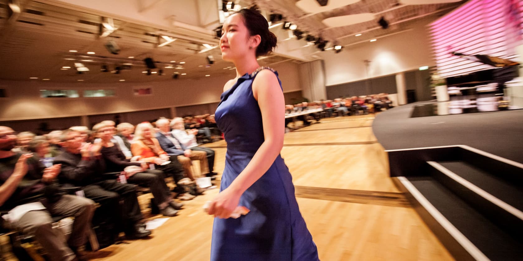 A woman in a blue dress walks on a stage in front of an audience.