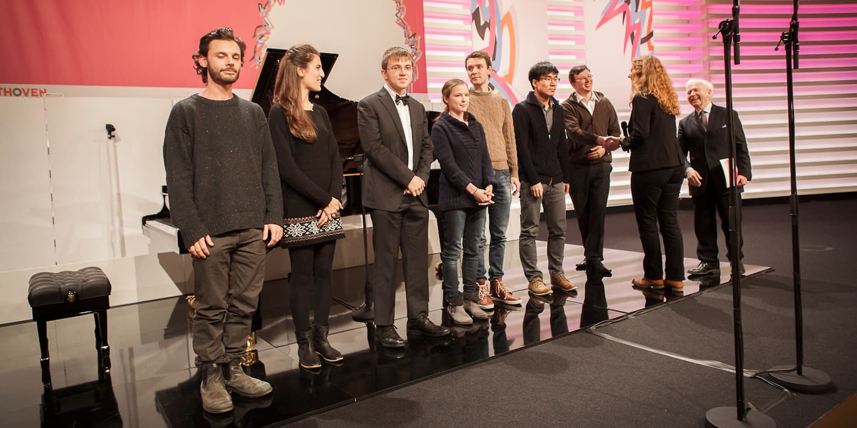 A group of people stands in a line on a stage. A woman shakes hands with a man. A grand piano is visible in the background.