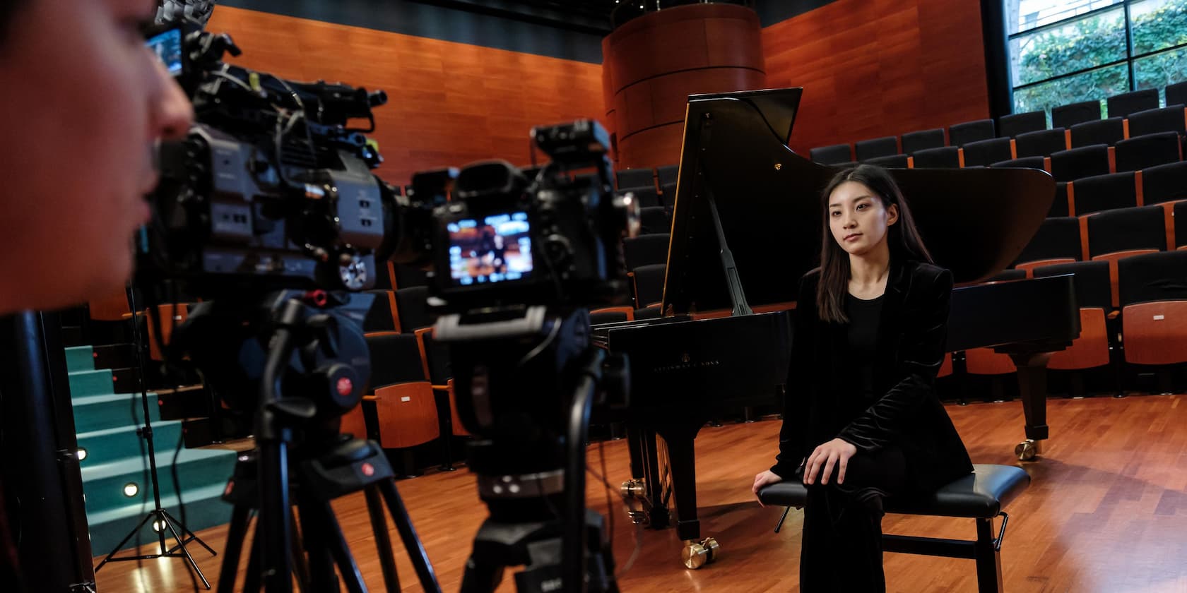 Woman sitting on a stage in front of a grand piano, being filmed by multiple cameras.