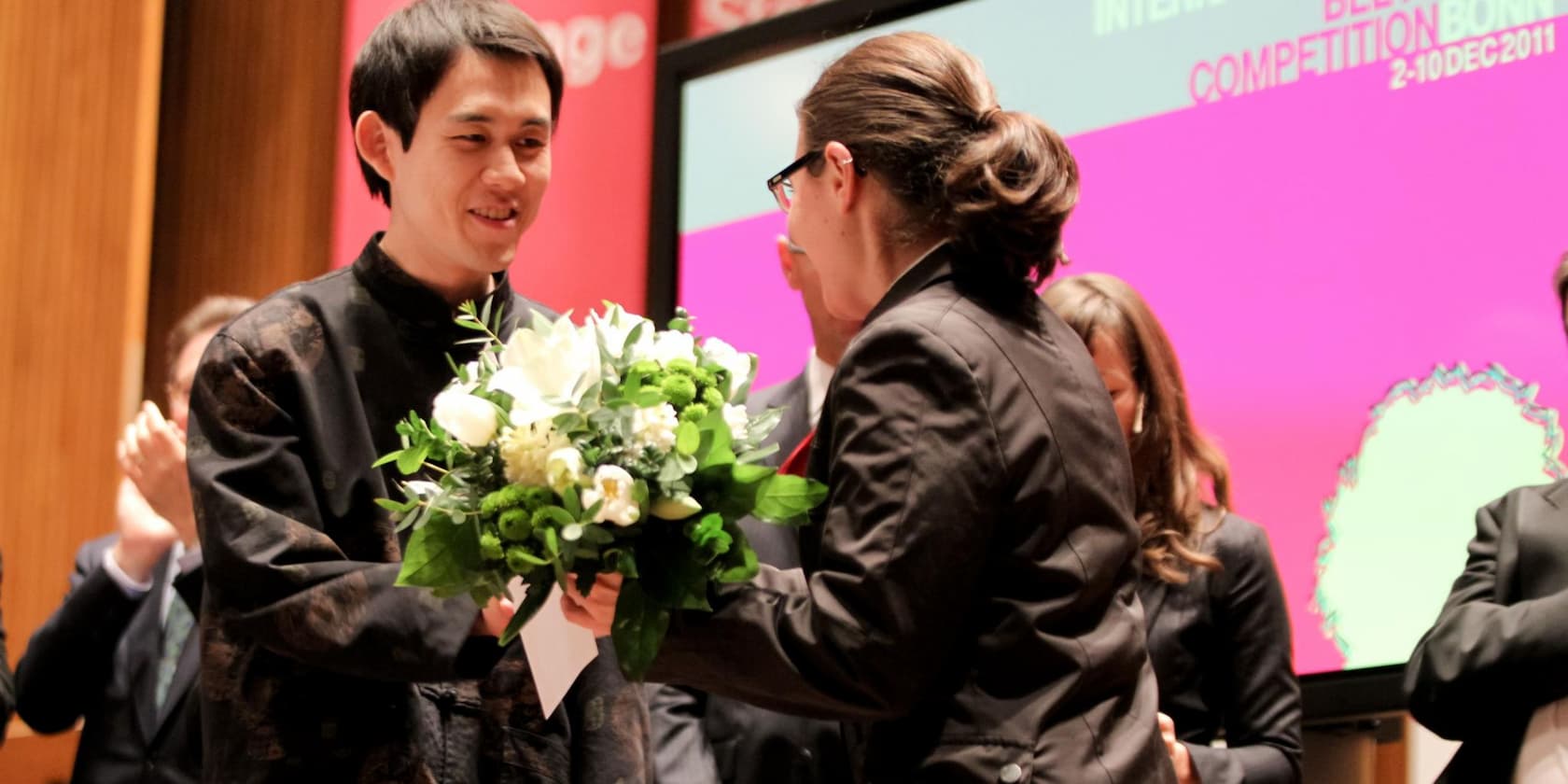 A person receiving a bouquet of flowers on stage while another person claps. In the background, there is a screen displaying the text 'INTERNATIONALE .... COMPETITION BONN 2-10DEC2011'.