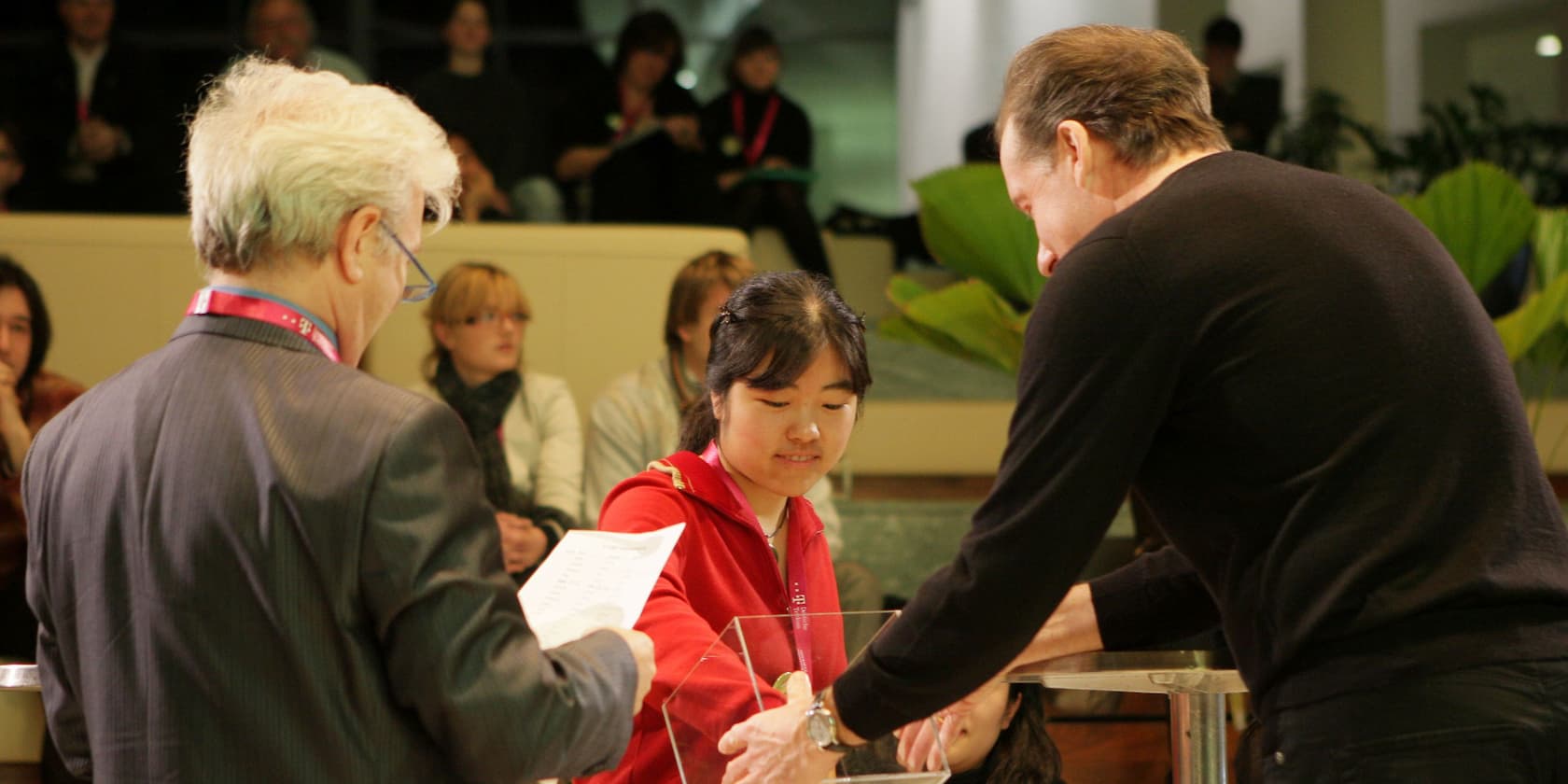 Eine Frau nimmt an einer Veranstaltung teil, bei der Preise oder Lose gezogen werden. Zwei Männer stehen daneben, einer hält ein Papier in der Hand. Im Hintergrund sitzen Zuschauer.