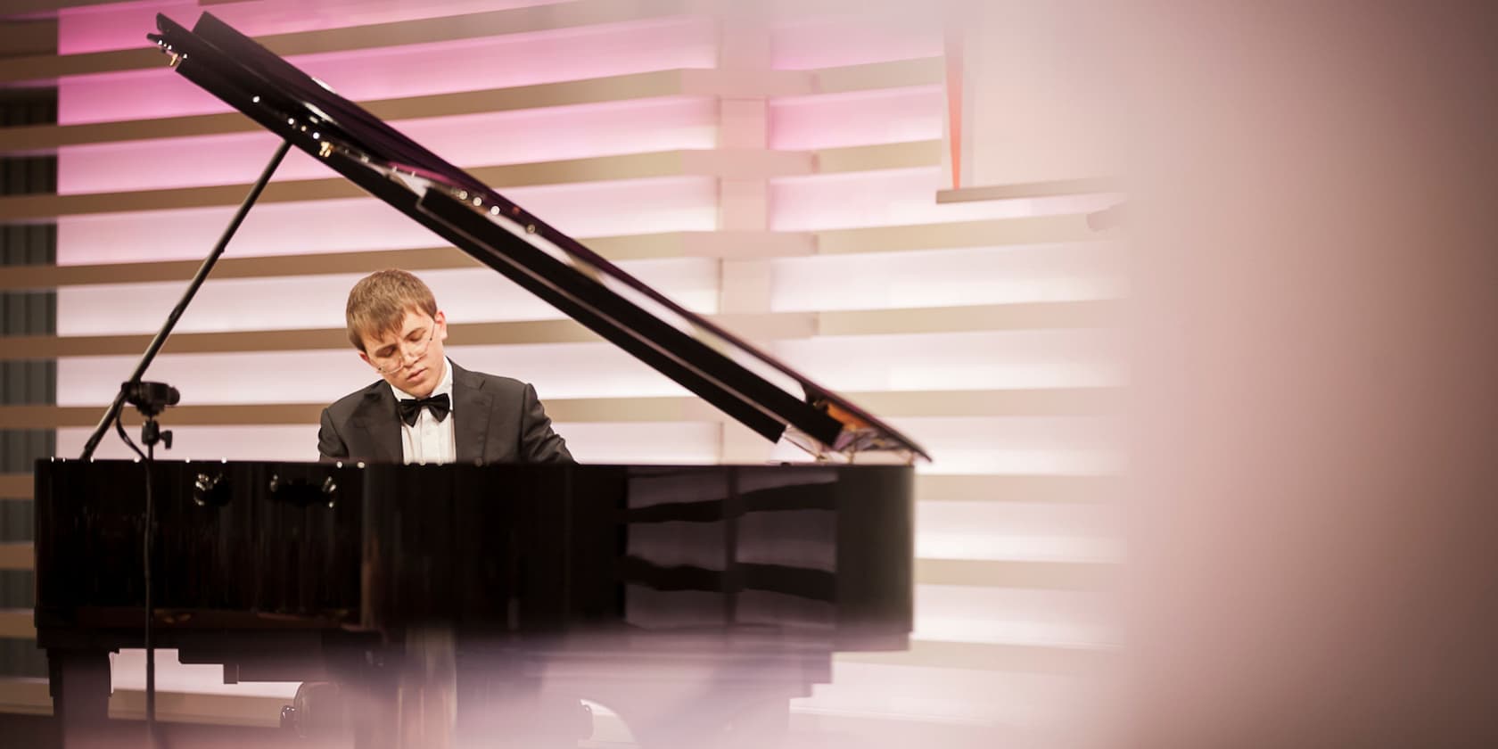 Man in a tuxedo playing piano on a stage with pink-lit walls.