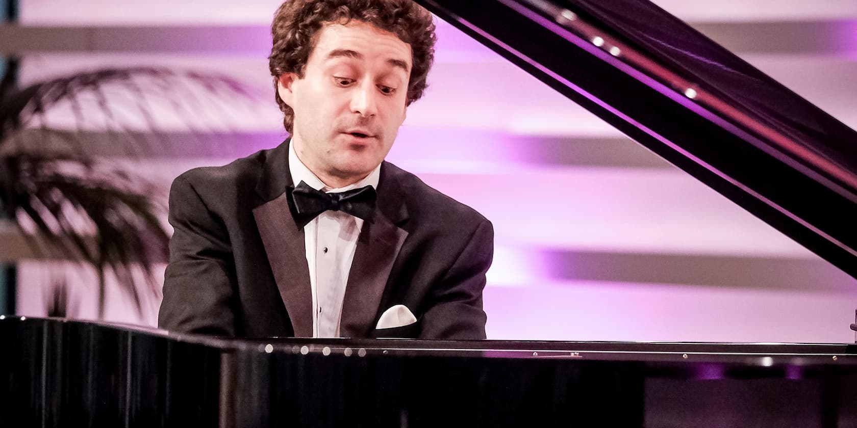 A man in a tuxedo playing the piano on a stage with purple lighting.