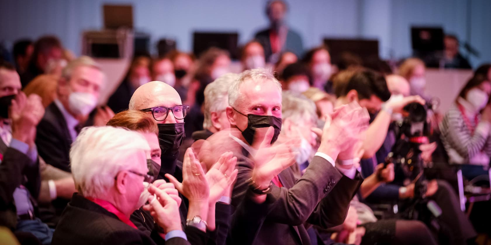 Crowd clapping during an event, many wearing masks.