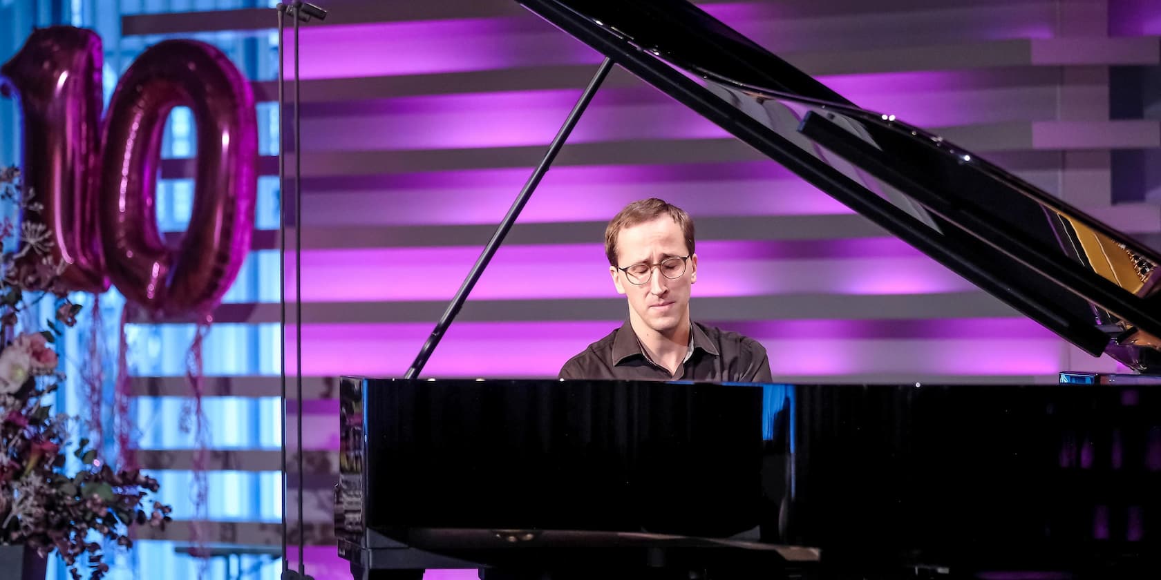 A man playing the piano at an event with purple lighting. In the background, there are large, pink balloons shaped as the number 10.