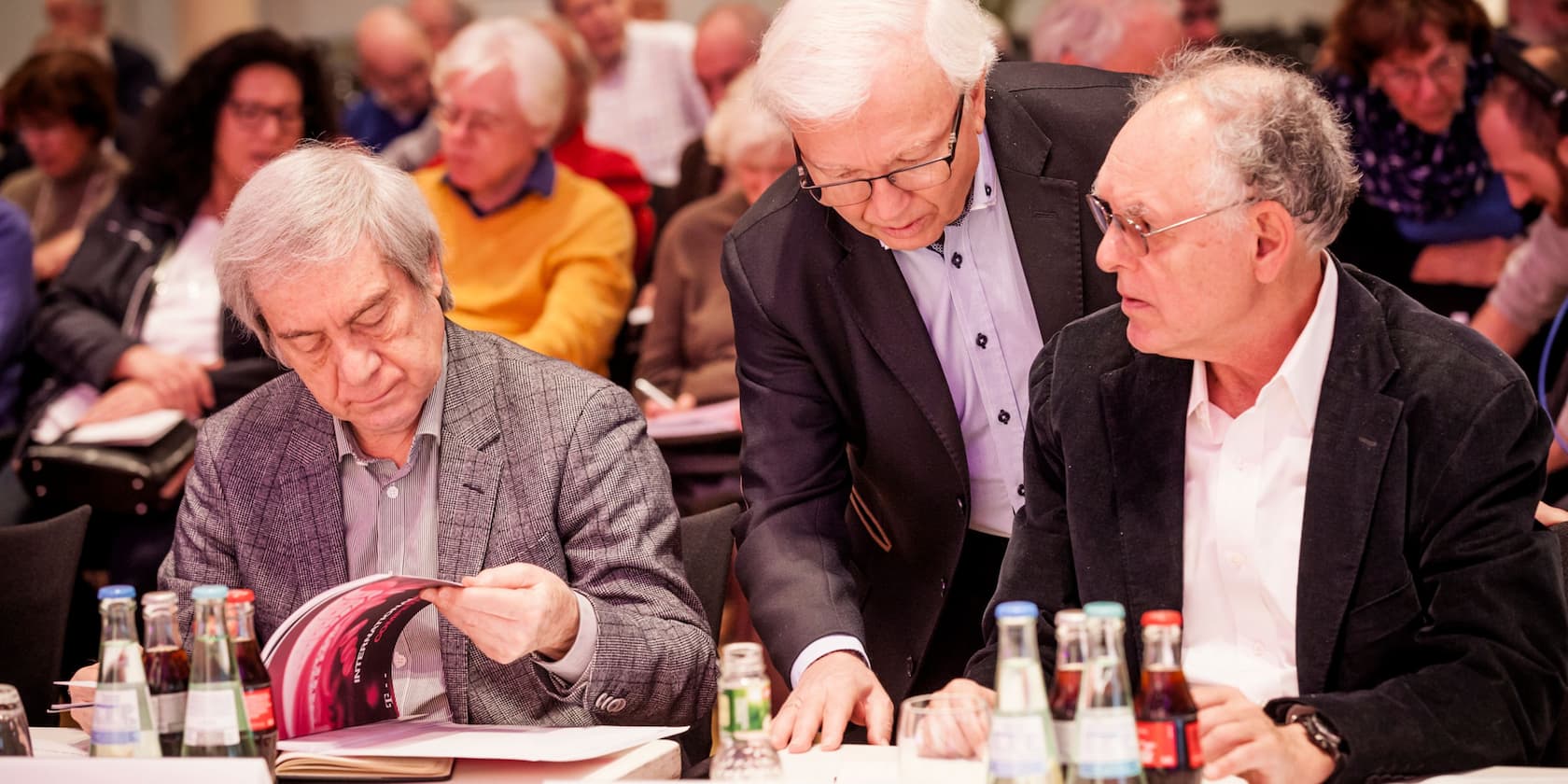 Elderly men discussing during a meeting, with other participants in the background.