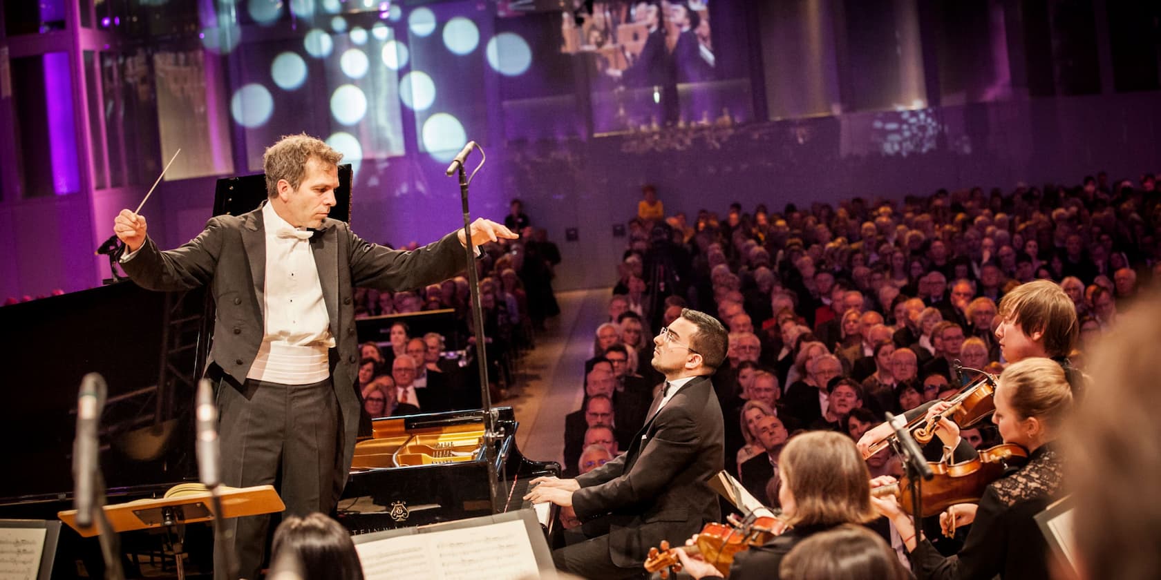 Dirigent leitet ein Orchester, während ein Pianist spielt, vor einem vollen Publikum in einem Konzertsaal.