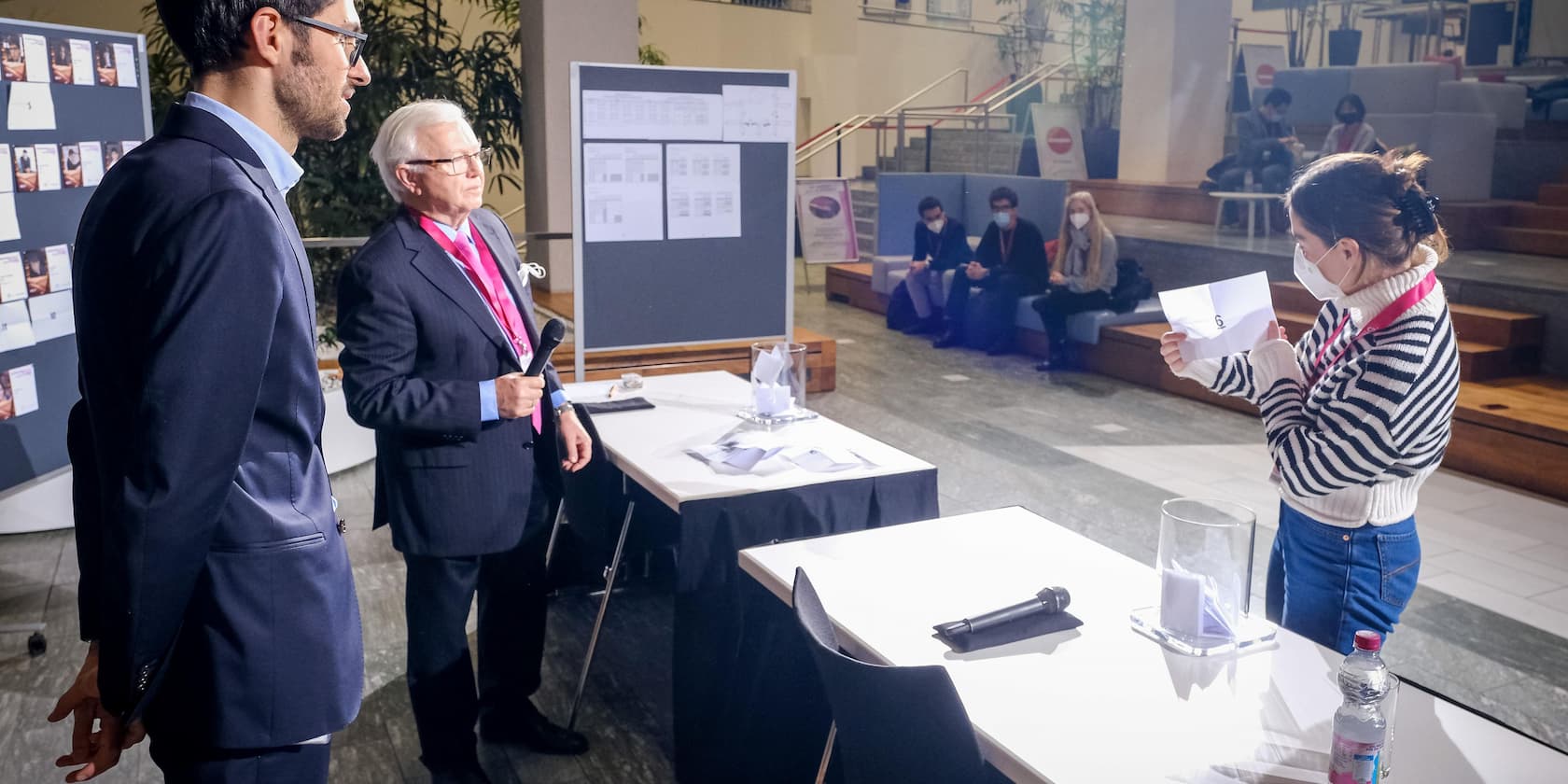 Zwei Männer und eine Frau, die eine Zeichnung zeigt, stehen in einem Raum mit Tischen und Posterboards. Im Hintergrund sitzen maskierte Menschen.