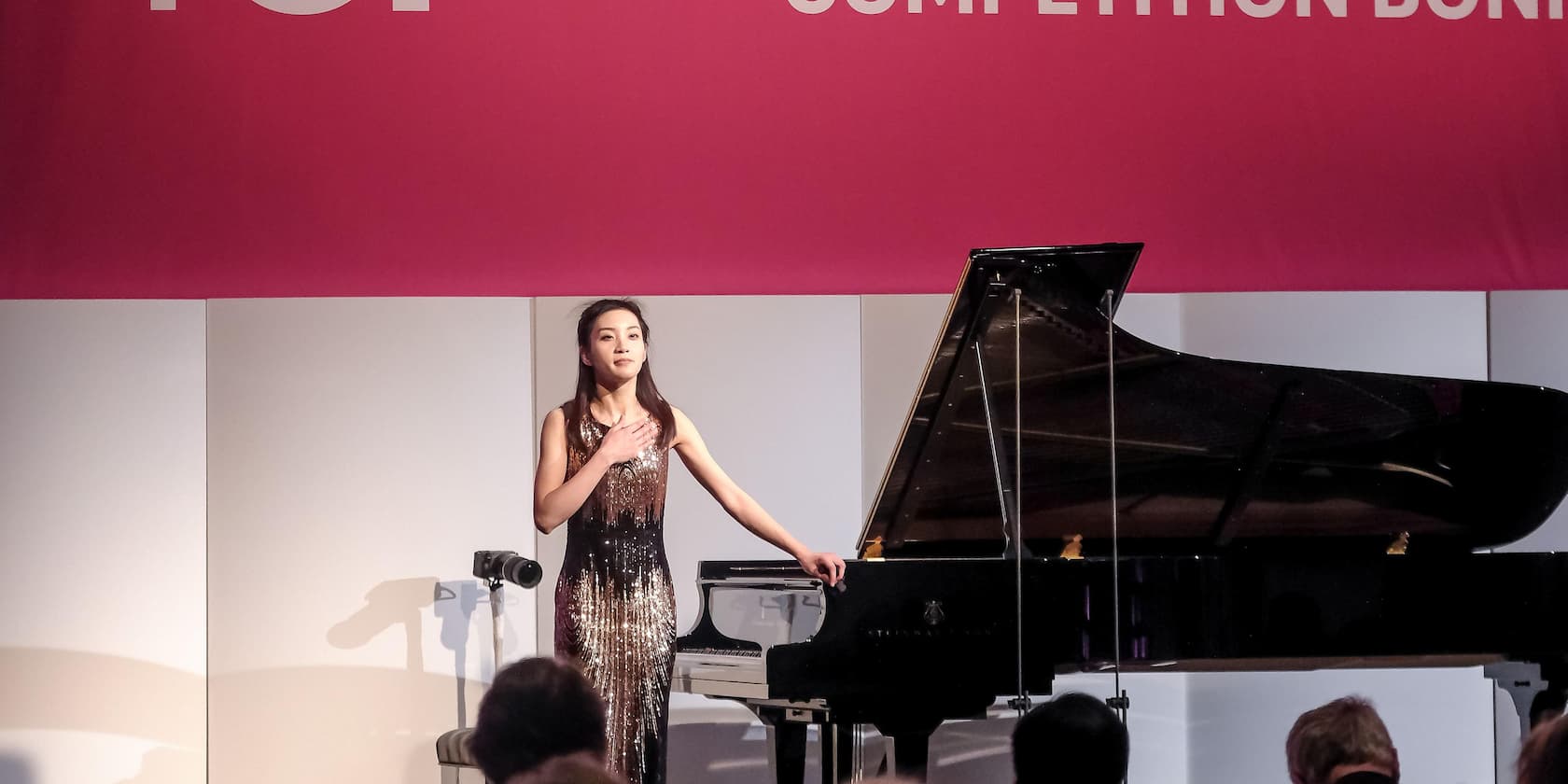 A woman stands next to a grand piano on a stage with one hand on her chest. In the background, a pink banner with the word 'COMPETITION' is visible.