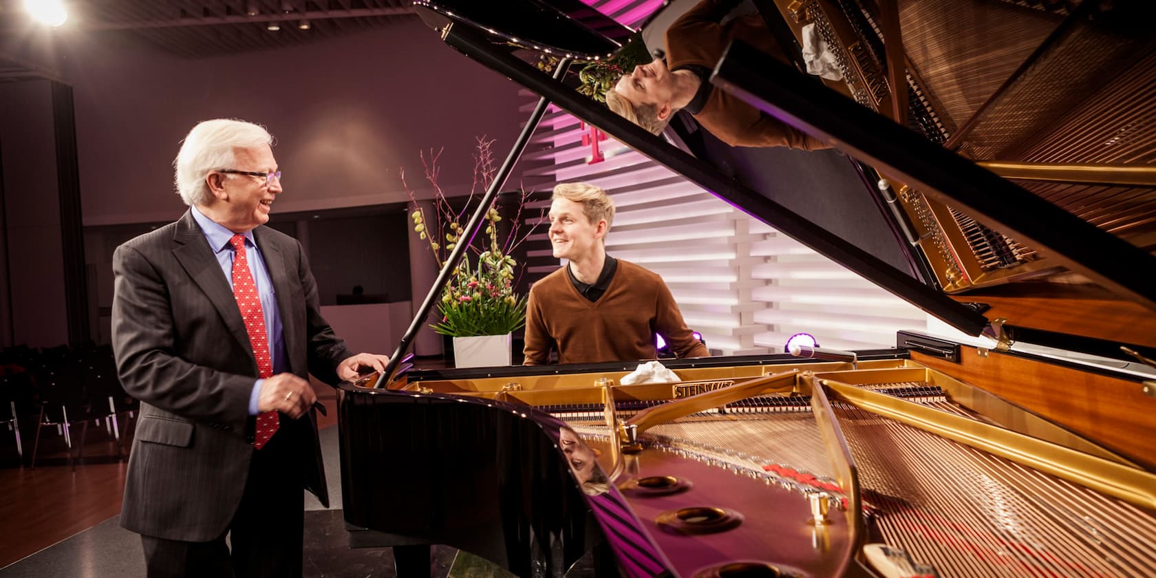 Older man in a suit talks to a younger man at a grand piano.