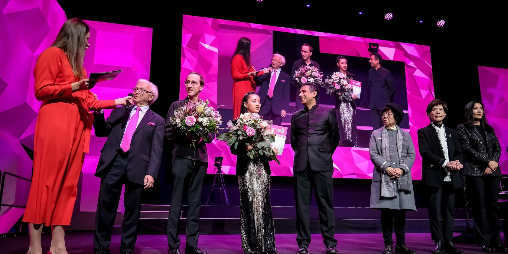 Several people on a stage at an award ceremony. Two are holding bouquets, and a woman is handing a microphone to an older man.