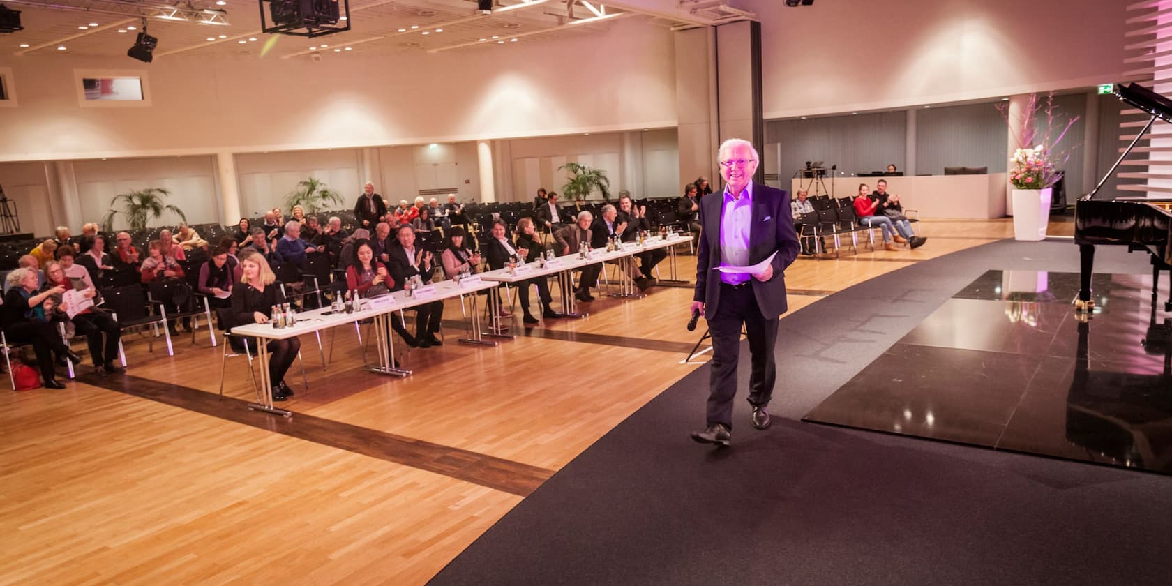 A man stands on a stage with a microphone, while an audience at tables applauds.