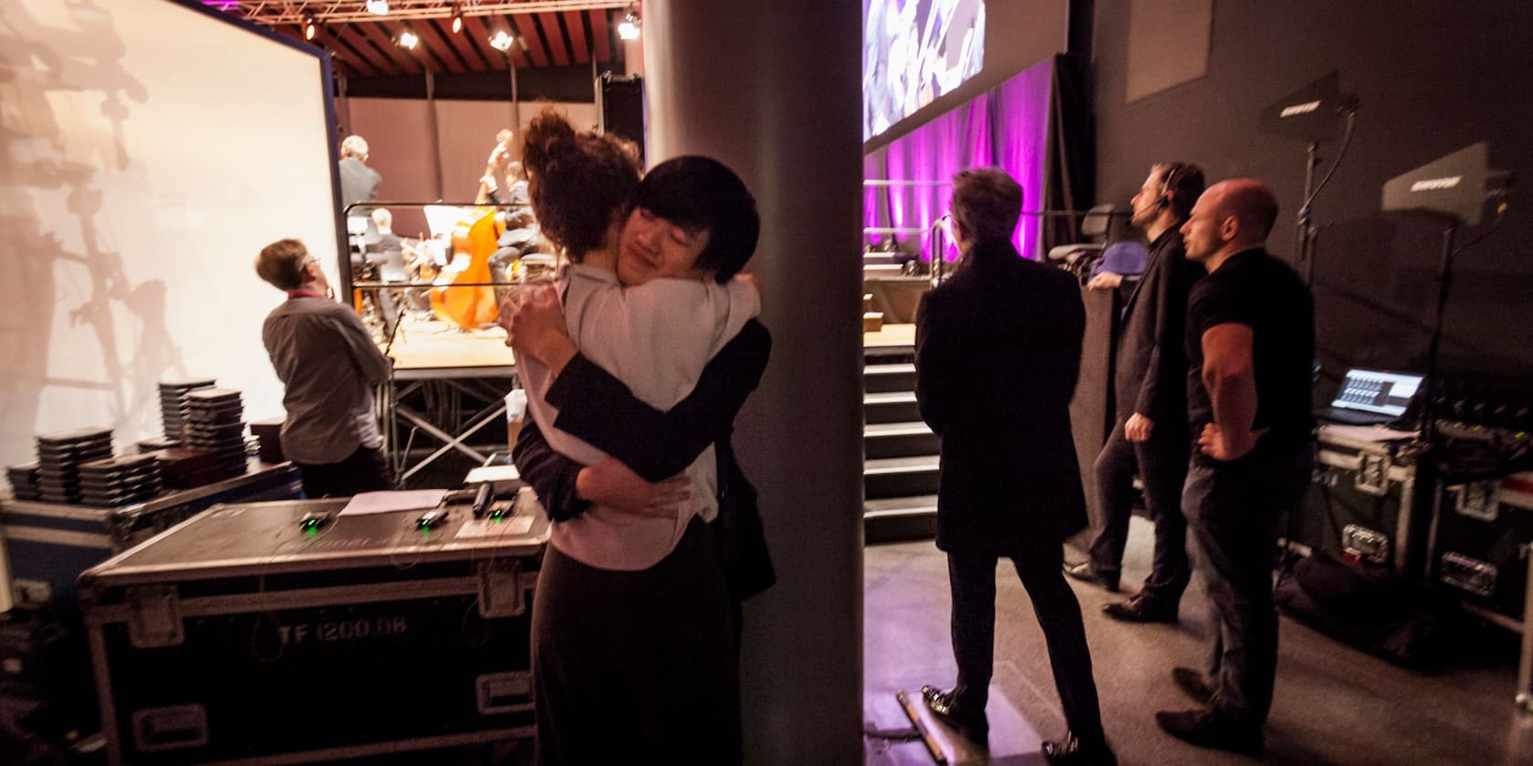 Two people embrace behind the stage of a concert while four others look on.