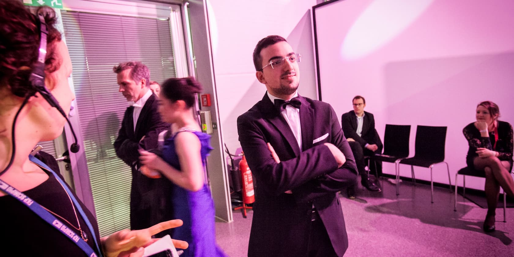 Several people backstage at an event. A man in a tuxedo stands with arms crossed while others are busy around him.