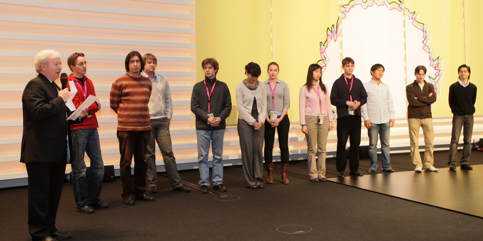 A group of people standing on a stage. A man on the left is speaking into a microphone.