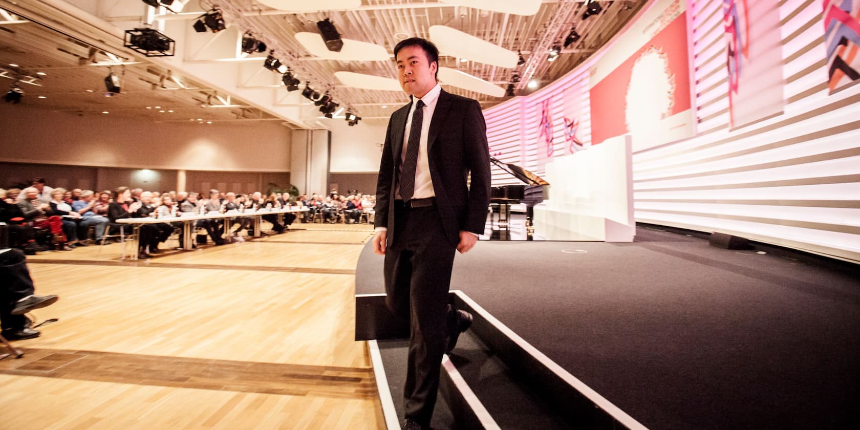 Man in a suit stands at the front of a conference room, with an audience sitting on chairs.