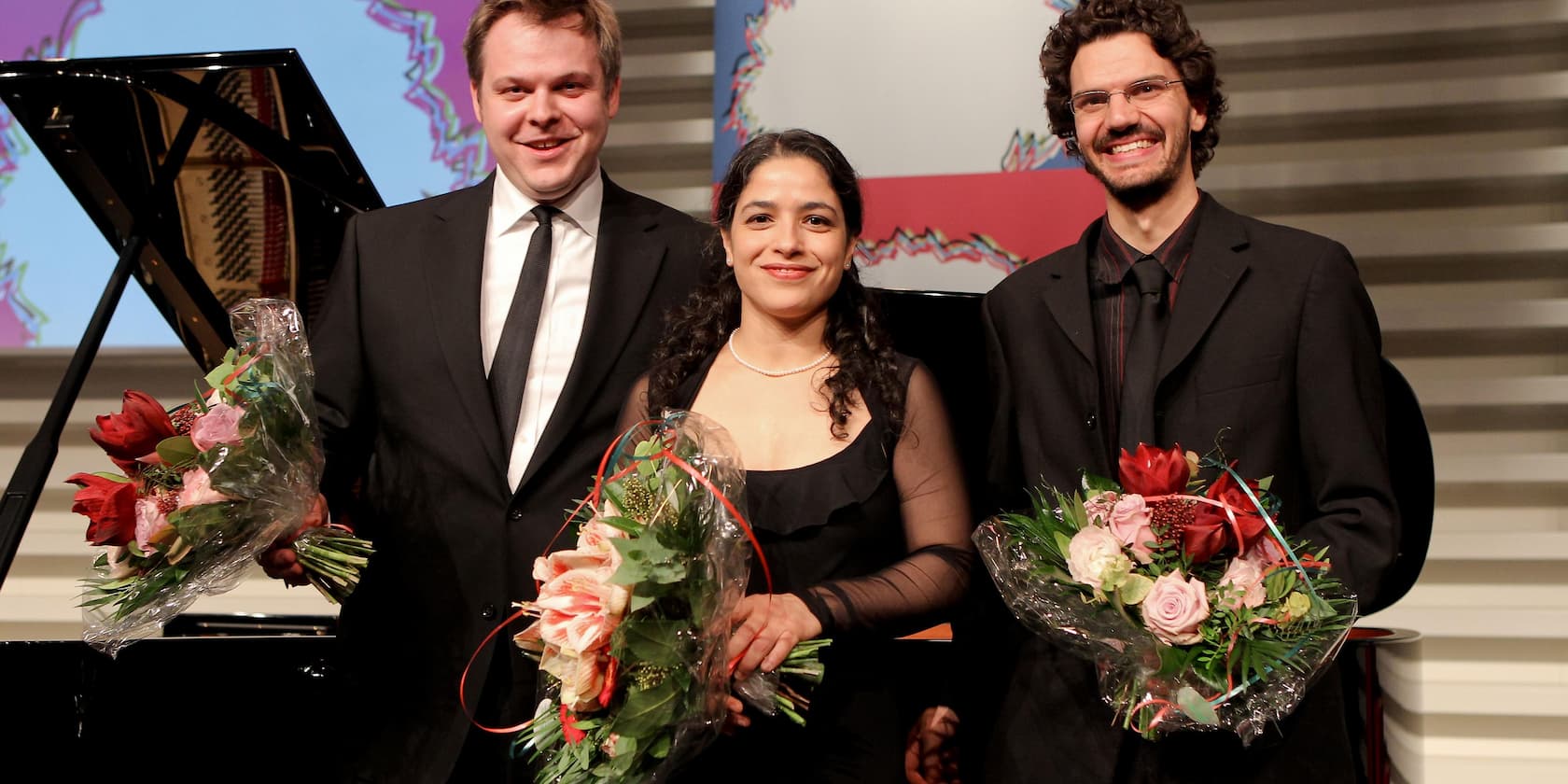 Drei lächelnde Personen in formeller Kleidung halten Blumensträuße in ihren Händen und stehen vor einem Klavier.