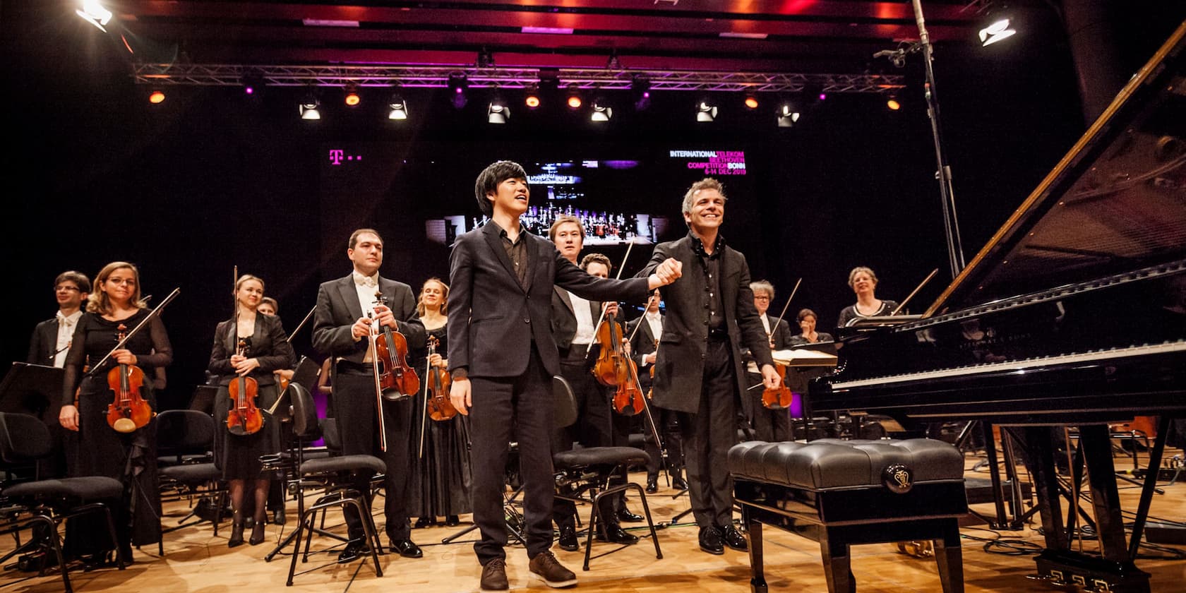 Musicians standing on stage after a concert, holding their instruments. A piano is seen in the foreground on the right.