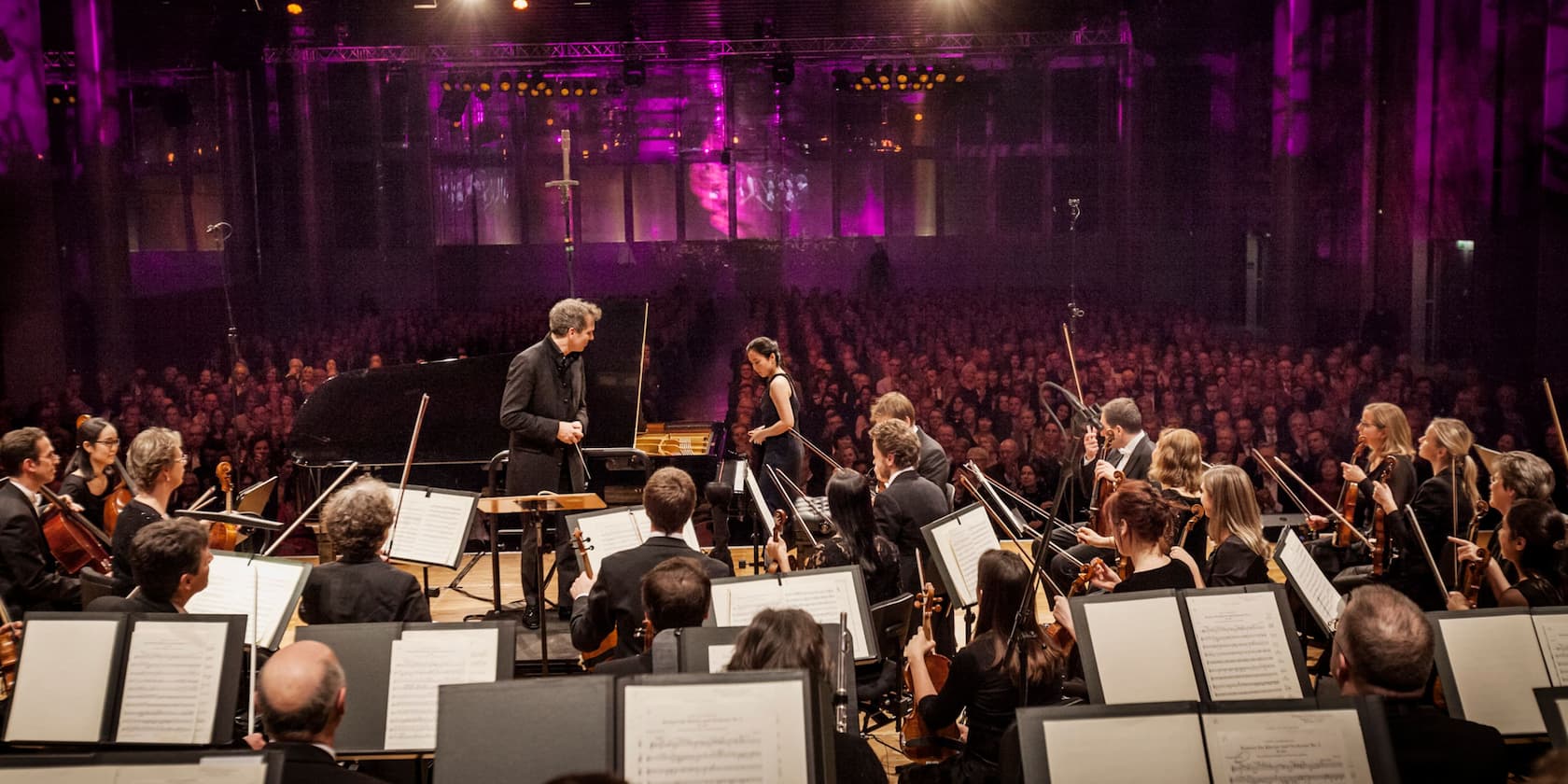 An orchestra performs in a concert hall with a full audience. In the foreground, musicians with instruments and music stands are visible.