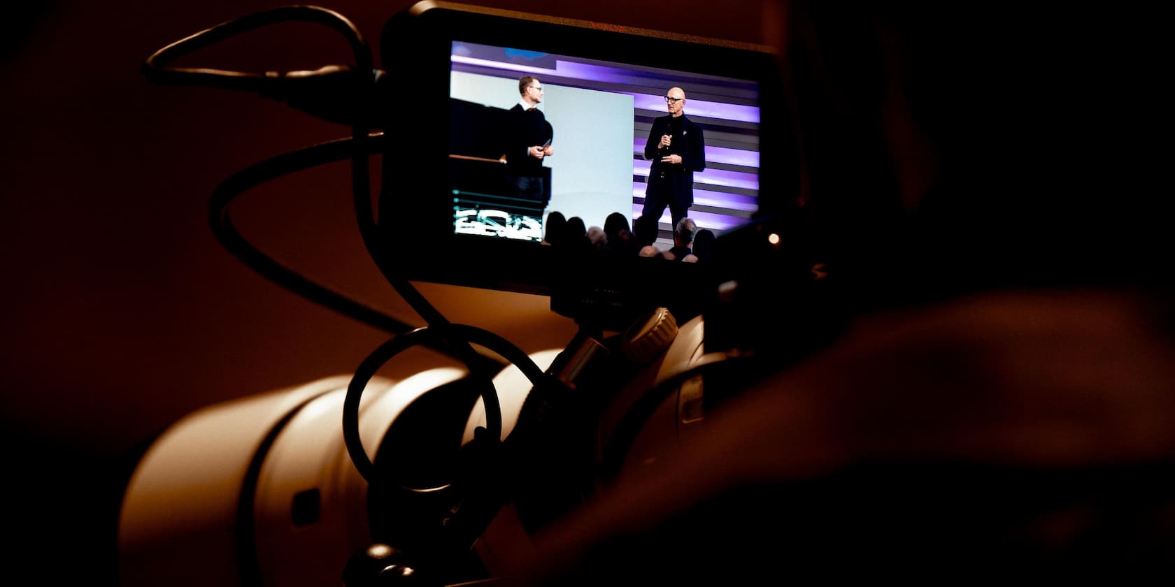 Two people are seen on a stage during a presentation on a camera screen.