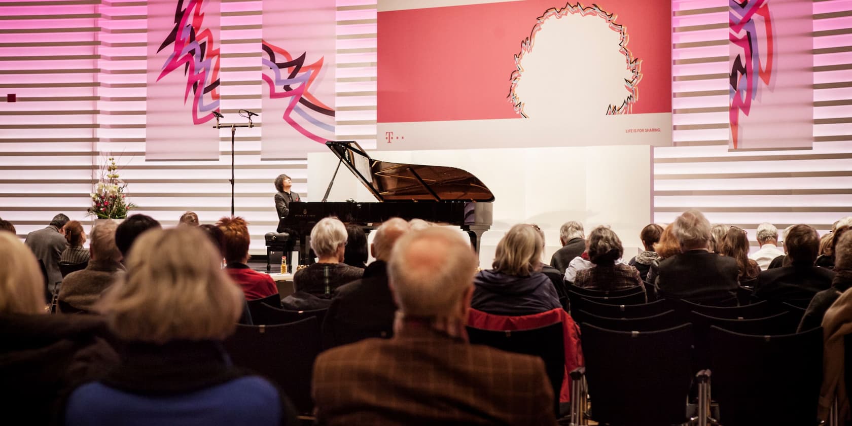 Ein Publikum sieht einem Konzert mit einem Flügel und einer Pianistin in einem modernen Saal zu.