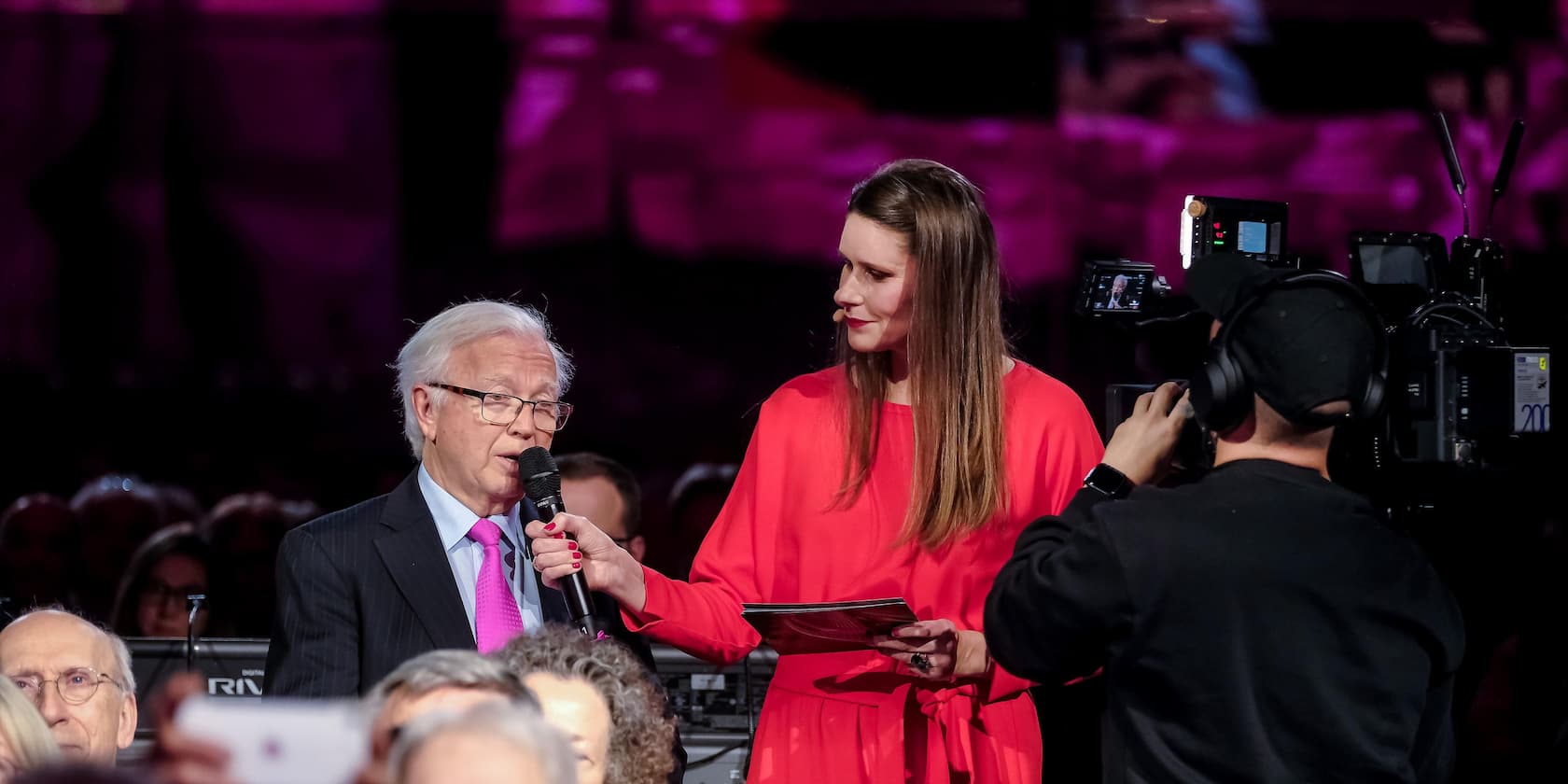 A moderator interviews an older man at an event while a cameraman films.